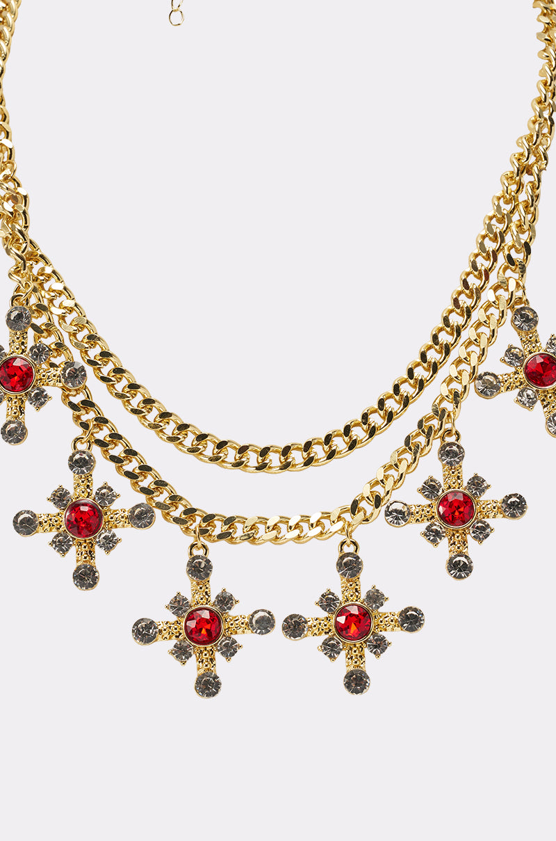 The MAXIMUM GLAM NECKLACE is a gold layered two-tier chain with six ornate cross crystal charms, each featuring sparkling silver stones and large red gemstones at the center, shown on a white background. 3
