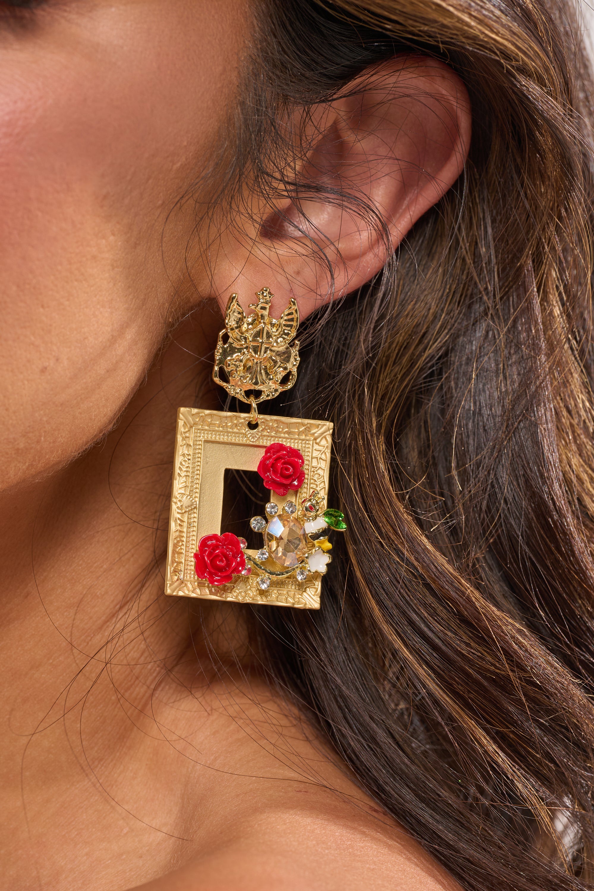 A close-up of a woman's ear wearing the PICTURE PERFECT EARRING, a large gold earring shaped like a decorative frame with red rose detail, small white flowers, green accents, and intricate top detailing. 1