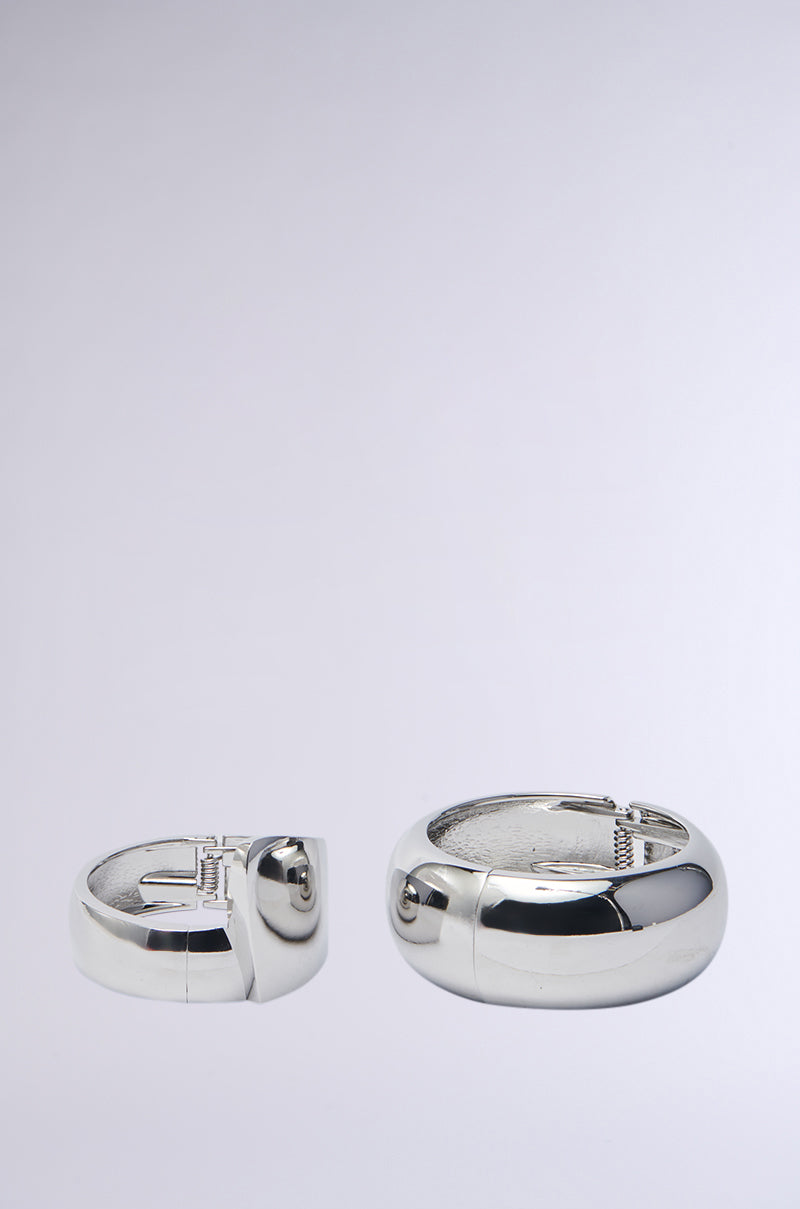 The ESSENTIAL BANGLE SET features two shiny faux silver bracelets on a plain gray background: a smaller flat bangle on the left and a larger rounder one on the right. Both have a sleek, reflective finish.