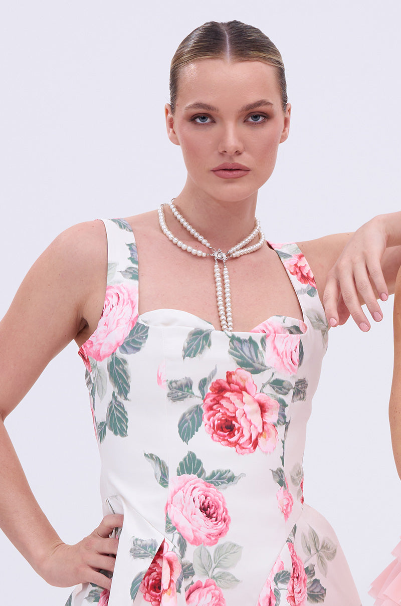A woman with slicked-back hair wears a white dress with pink florals and a draped design, styled with the DRIPPIN PEARLZ NECKLACE, posing with one hand on her hip against a plain light background.