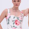 A woman with slicked-back hair wears a white dress with pink florals and a draped design, styled with the DRIPPIN PEARLZ NECKLACE, posing with one hand on her hip against a plain light background.