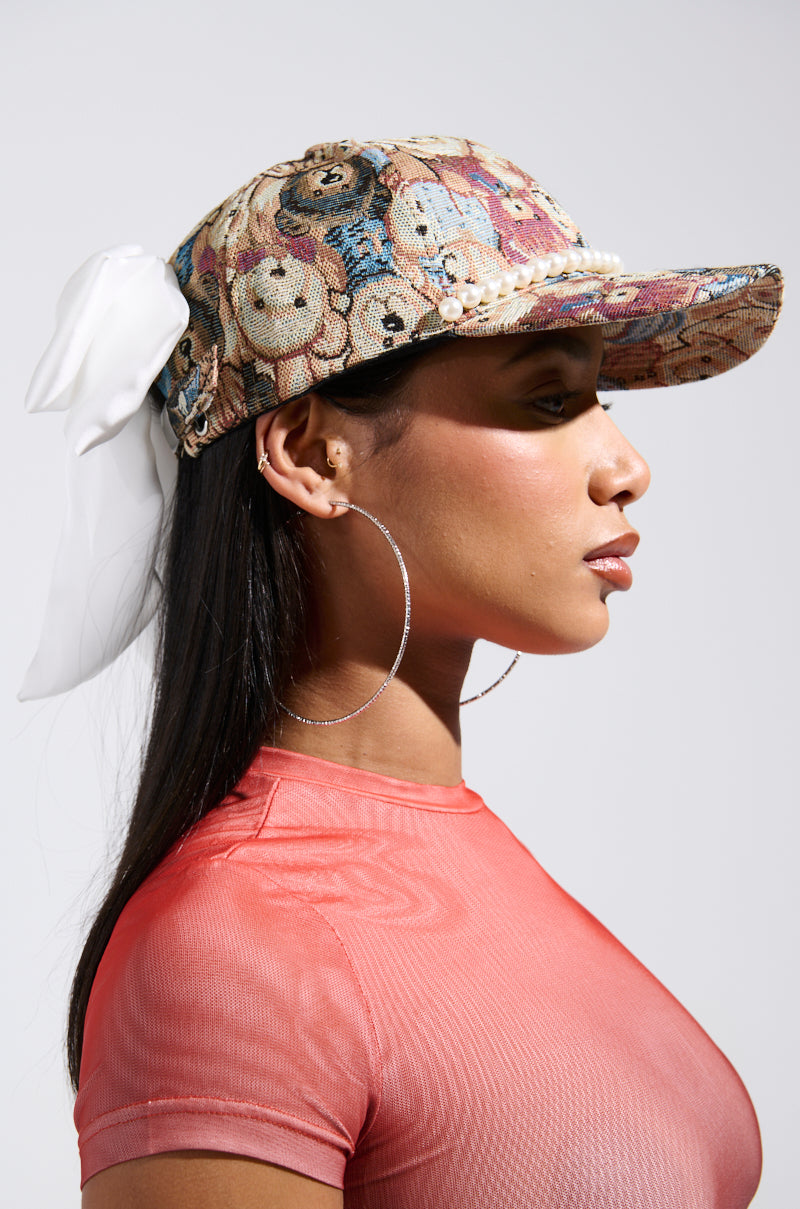 A woman wears the BEARY NICE BOW HAT with a delicate silk bow at the back, complemented by a sheer pink top and large hoop earrings, standing against a plain background.