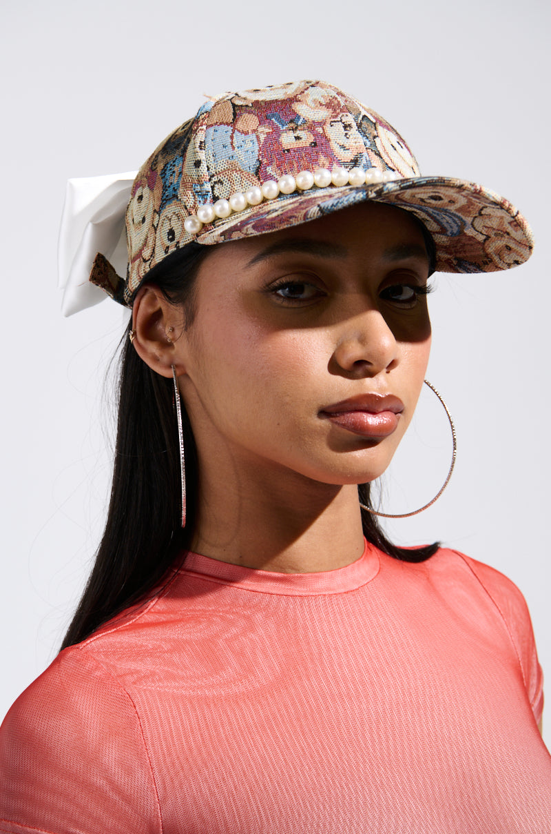 Wearing the BEARY NICE BOW HAT, adorned with a colorful bear print and a silk bow at the back, she gazes confidently. Her long hair complements the artistic pattern as large hoop earrings dangle beside her sheer pink top.