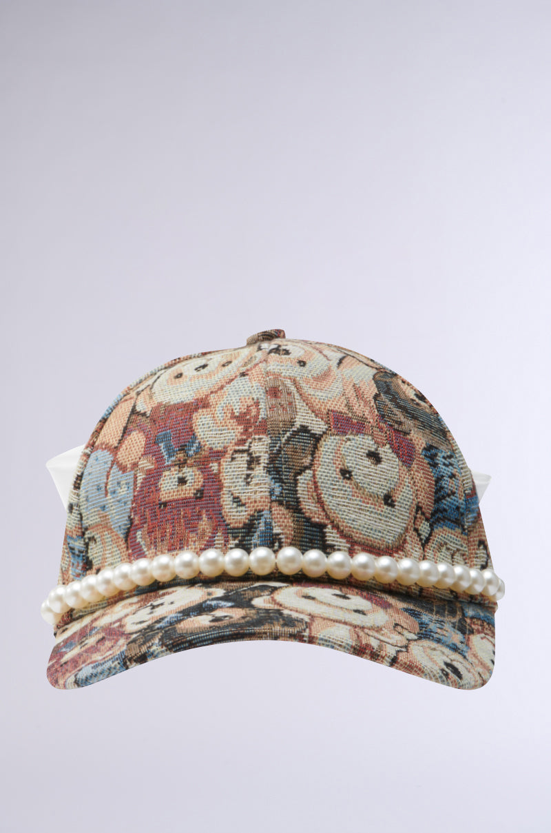 The BEARY NICE BOW HAT is a baseball cap with a beige cotton tapestry design showcasing patterns and characters. Faux pearls adorn the front edge, while white fabric points at the back resemble a crown or ears. A plain gray background enhances its charm.