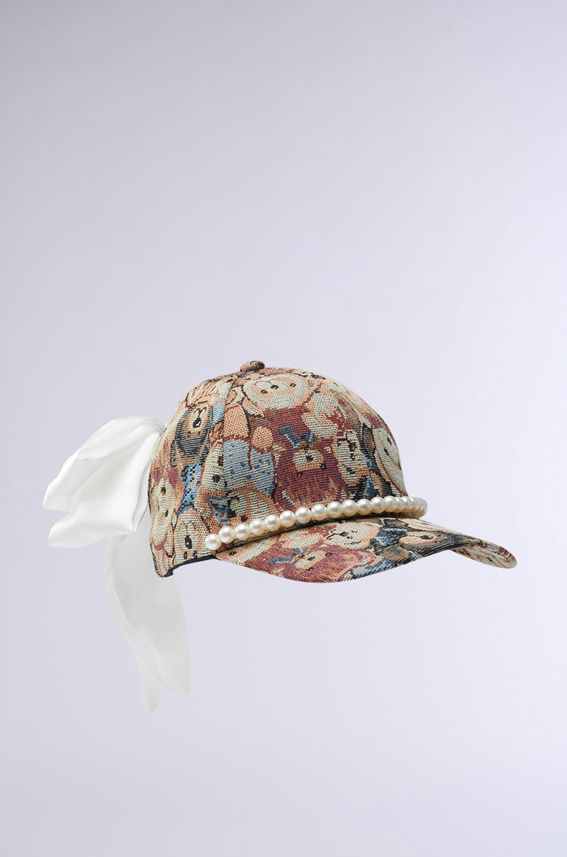 The BEARY NICE BOW HAT is a cotton tapestry baseball cap adorned with various dog faces, featuring a brim embellished with pearls and a large white silk bow at the back, set against a plain light backdrop.