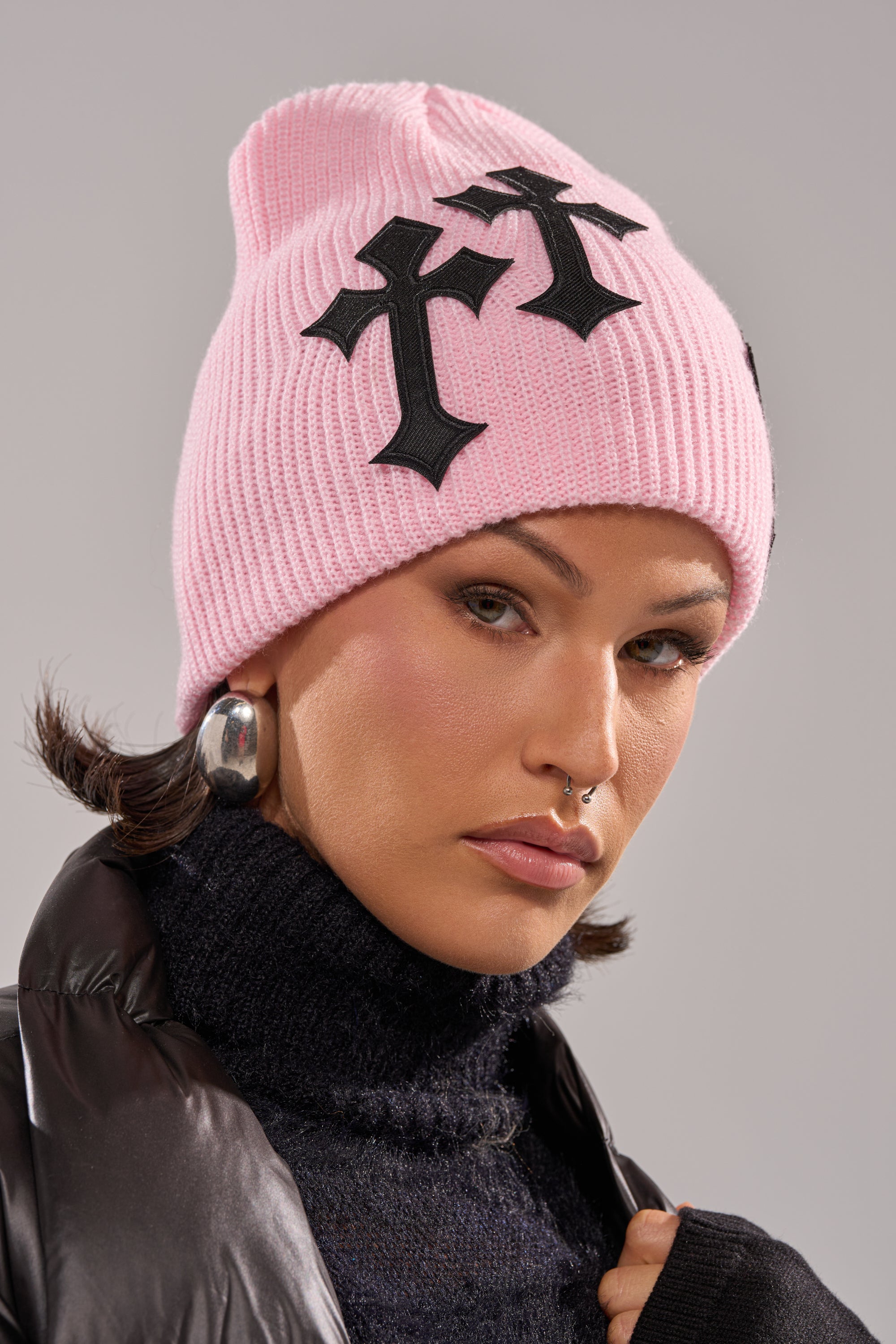 A person wears the CROSS ME BEANIE in baby pink with black crosses, silver hoop earrings, a black turtleneck, and a black puffer jacket, looking confidently at the camera.