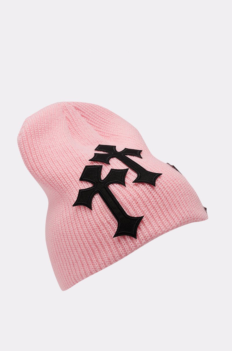 The CROSS ME BEANIE is a baby pink ribbed knit beanie with three large black gothic-style cross patches on the front, shown against a plain white background. 2