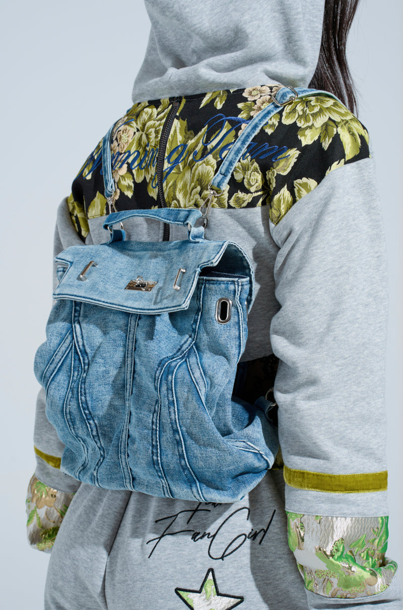A person in a gray hoodie with floral and green accents that says "Femi Girl" on the back carries the KEEP EM ALL BACKPACK—a blue denim bag with decorative stitching, silver hardware, and adjustable straps for versatile style. 2