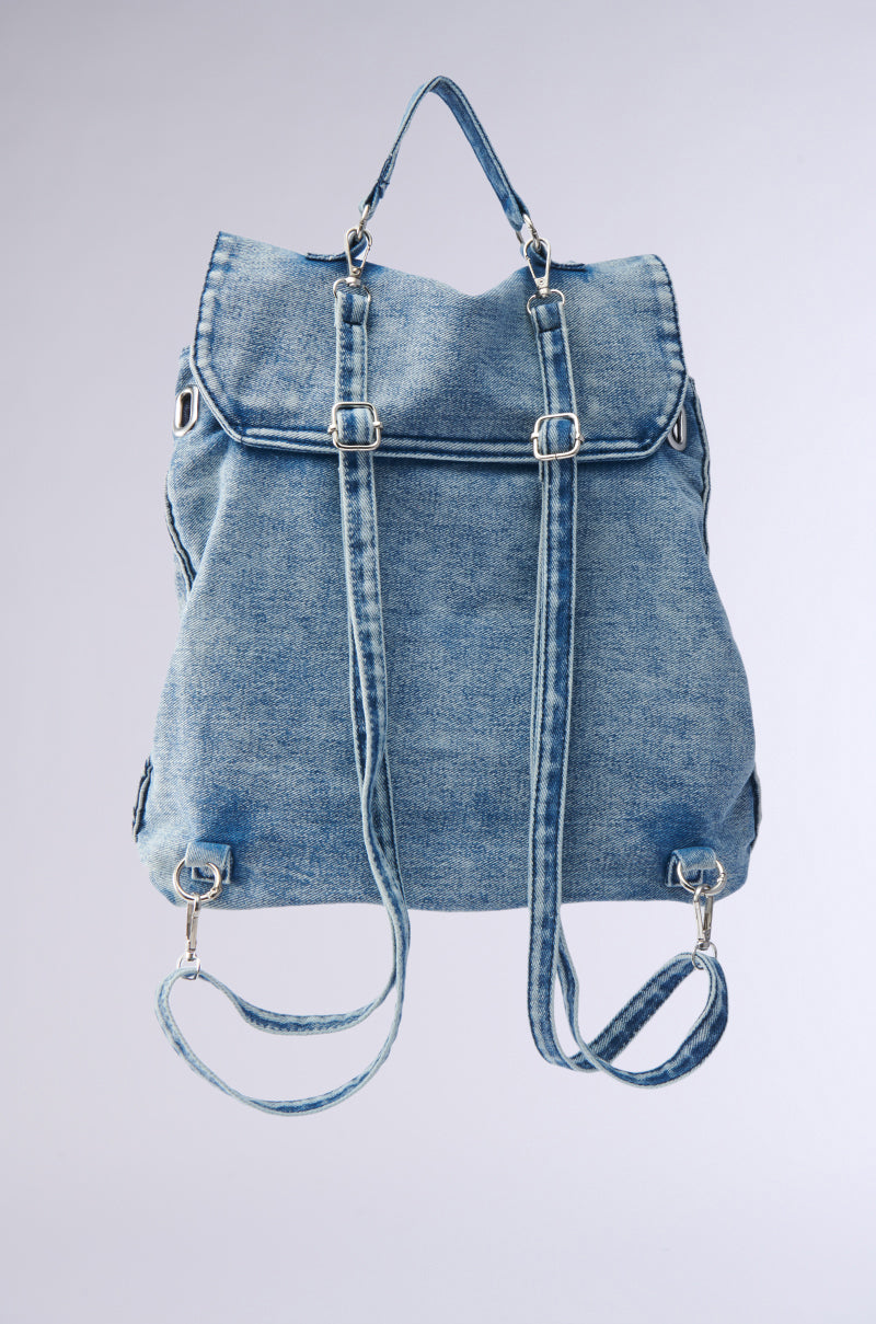 The KEEP EM ALL BACKPACK is a light blue denim bag with silver buckles, adjustable shoulder straps, a top handle, and flap closure. This versatile accessory stands out against a simple, light background. 3