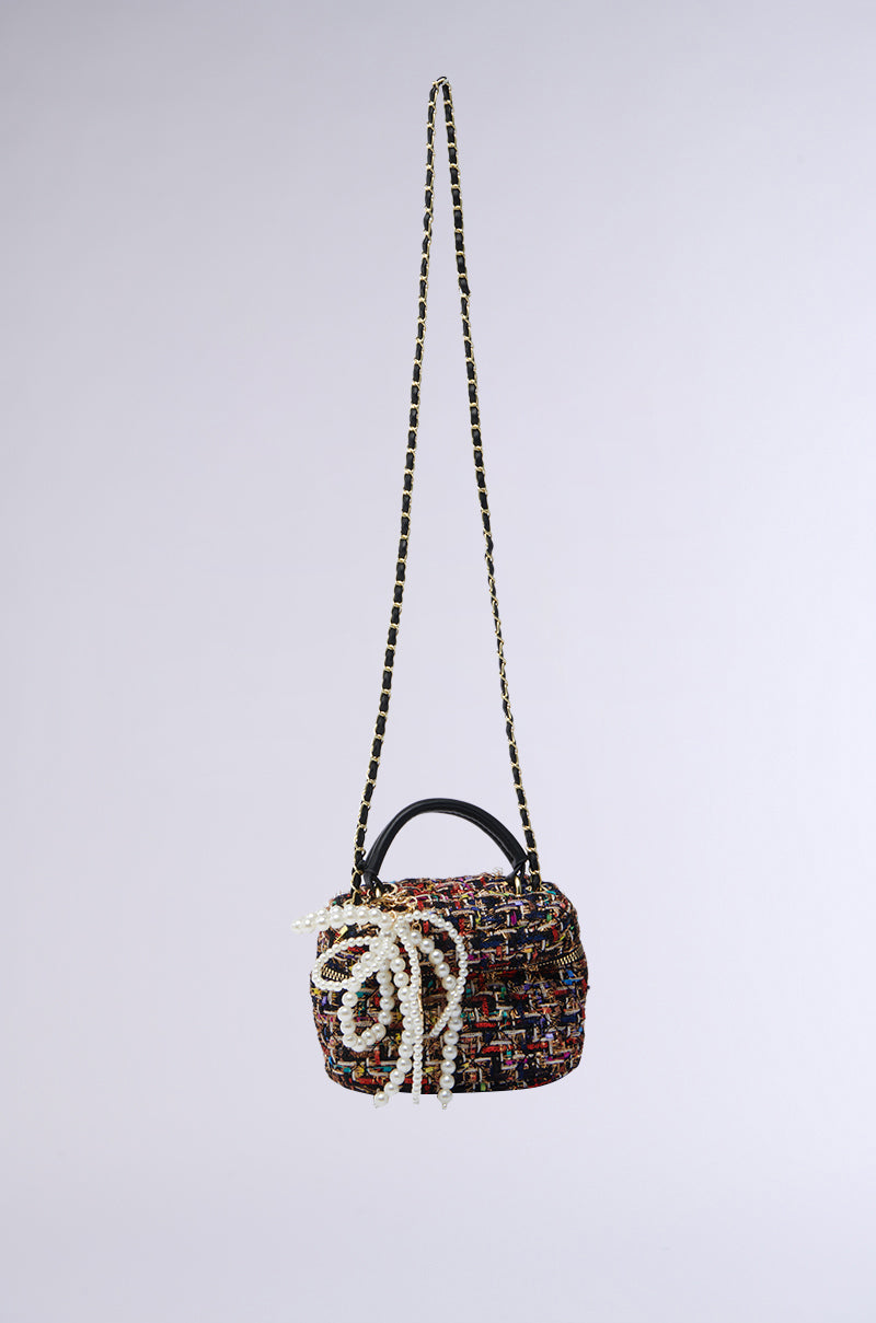 The IN THE CITY MINI JUNK BAG, a multicolored woven handbag crafted from quilted tweed, floats in mid-air. It has a long strap and small top handle, accented with decorative pearl bow charms on the front against a plain light grey background.