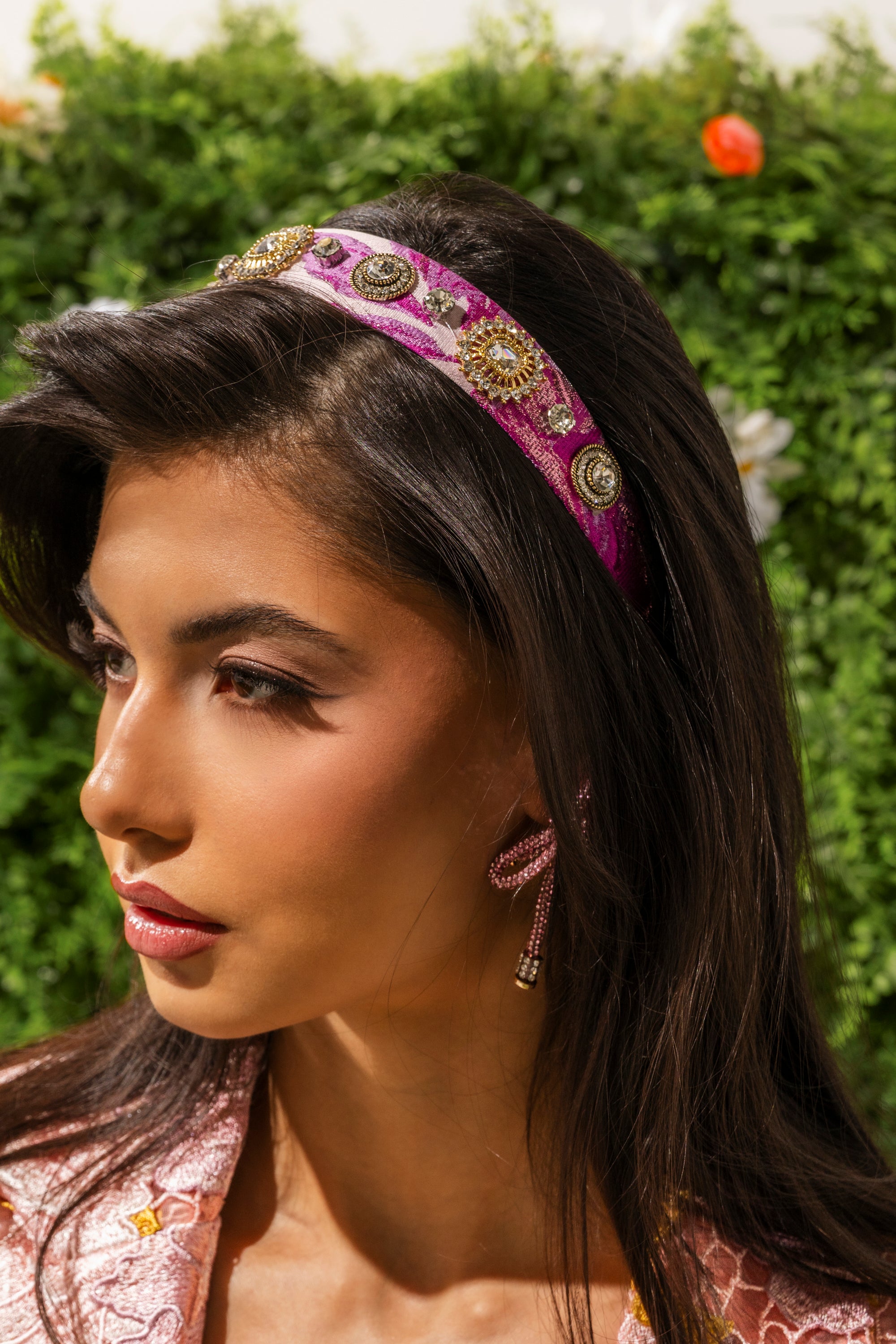 A woman with long brown hair wears the GIRLS GIRL EMBELLISHED HEADBAND and a matching earring. She has soft makeup with winged eyeliner and pink lipstick, set against green foliage with orange and white flowers. 0