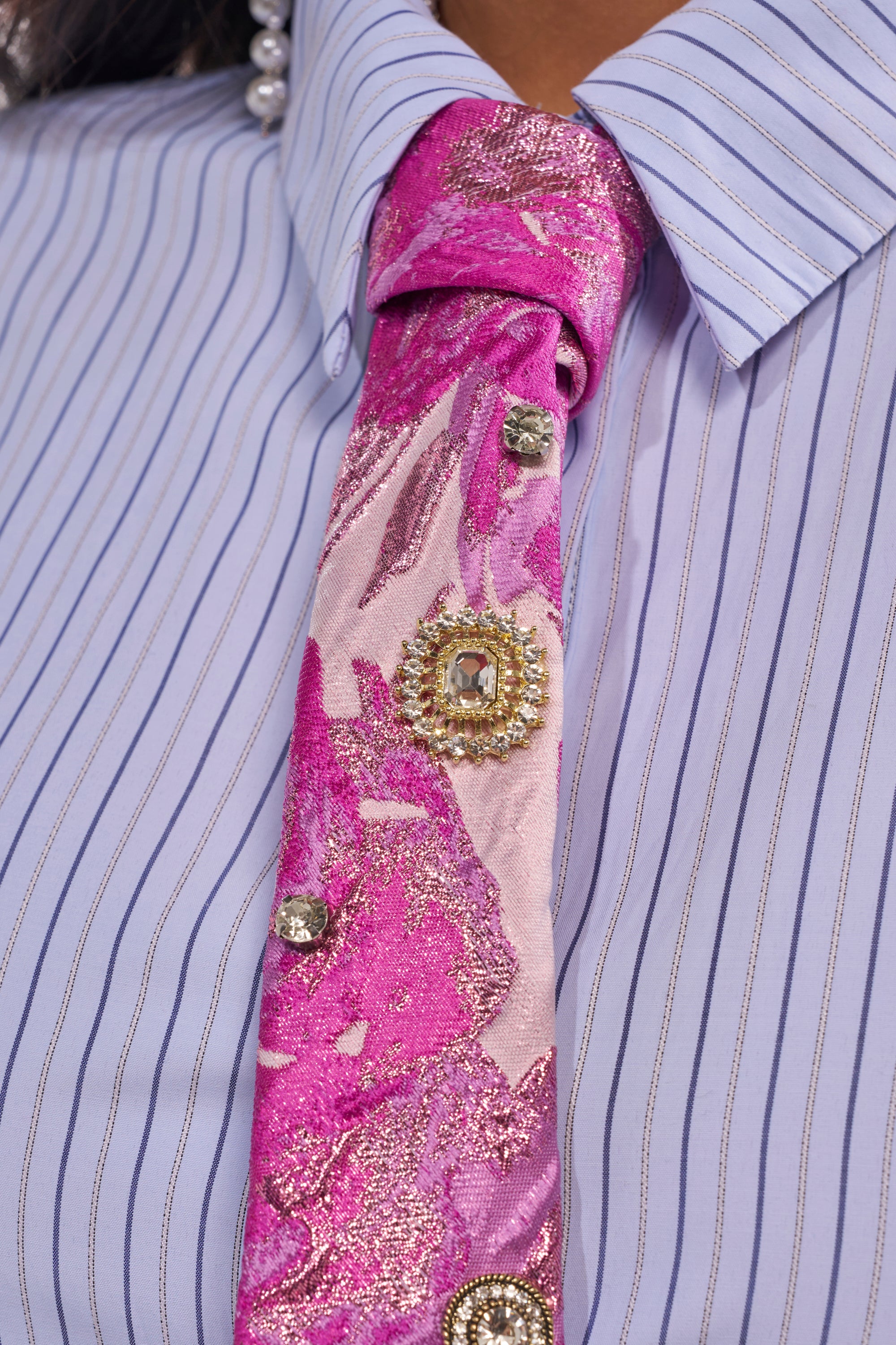 Close-up of a person wearing the GIRLS GIRL EMBELLISHED TIE in pink with glittery patterns and jeweled brooches, paired with a blue and white pinstripe collared shirt. A pearl earring is partially visible. 2
