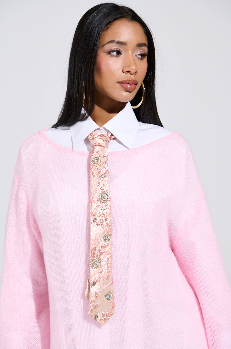 A woman with long dark hair wears a light pink sweater over a white collared shirt, accented by the LIVE FOR IT EMBELLISHED TIE featuring a shimmering metallic floral pattern. Hoop earrings complete her look as she gazes to her left against a plain background.