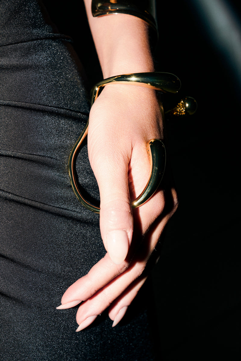 A hand with long, manicured nails rests on black fabric, showcasing the YOU COULD NEVER BRACELET—a sculptural piece that wraps around the wrist and thumb for a bold statement look.