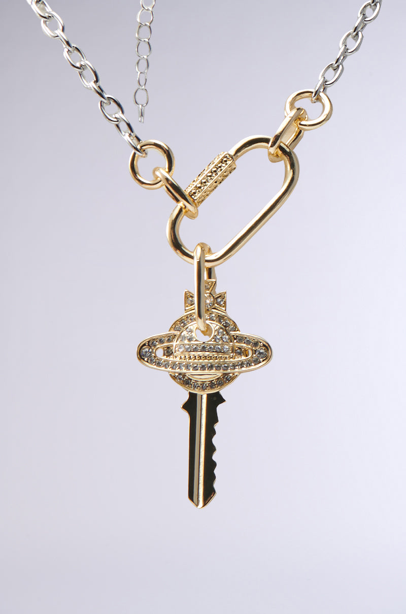 The TAKE ME AWAY NECKLACE is a gold and silver chunky piece featuring a sparkling gold key pendant with a circular rhinestone-encrusted emblem, attached by a carabiner centerpiece and displayed against a plain background. 2