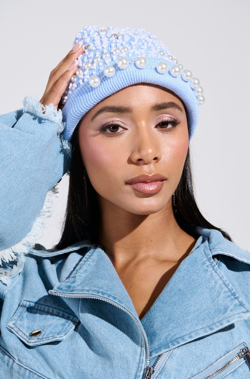 A woman touches her head while wearing the IT GURL EMBELLISHED BEANIE adorned with pearlescent beads and rhinestones. She's dressed in a denim jacket with frayed edges against a plain, light-colored background.