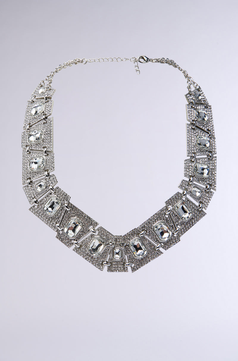 The CLEO ICED OUT NECKLACE is a silver chain link necklace with rectangular, faceted clear gemstones set in ornate, textured geometric plates, shown on a plain light gray background.