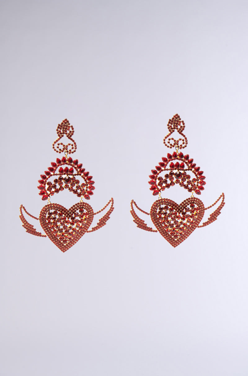 The CATCH MY HEART EARRING features ornate, dangling heart-shaped designs in red and gold. With bold winged accents and intricate floral detailing, they exude a flaming heart motif against a simple white background. 1