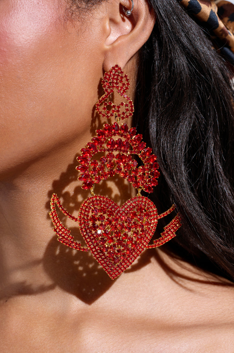 Close-up of a person wearing CATCH MY HEART EARRING, adorned with red rhinestone embellishments and sparkling stones. These earrings feature intricate details, hanging elegantly against dark hair. 2