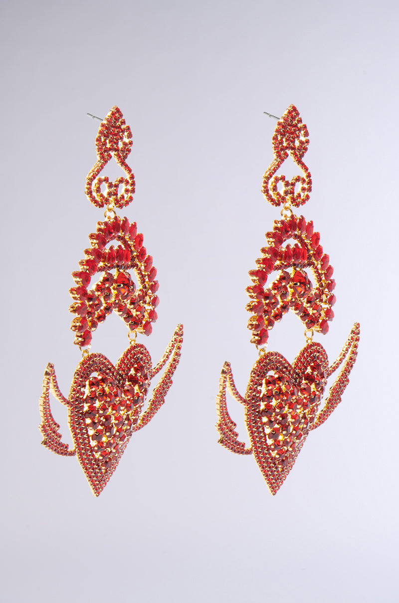 The CATCH MY HEART EARRING showcases intricate flaming heart designs in red and gold, with bold winged accents. Sparkling gemstones and reflective surfaces make the red rhinestone embellishments pop against a plain background. 3