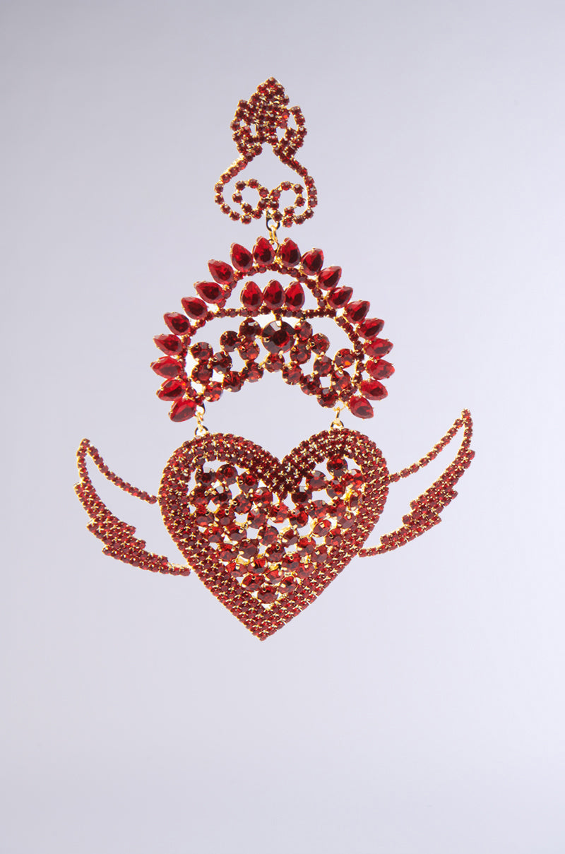 The CATCH MY HEART EARRING features an elaborate red heart with bold winged accents and red rhinestones, suspended against a simple gray background. It showcases intricate details, including wing-like extensions and decorative loops above. 4