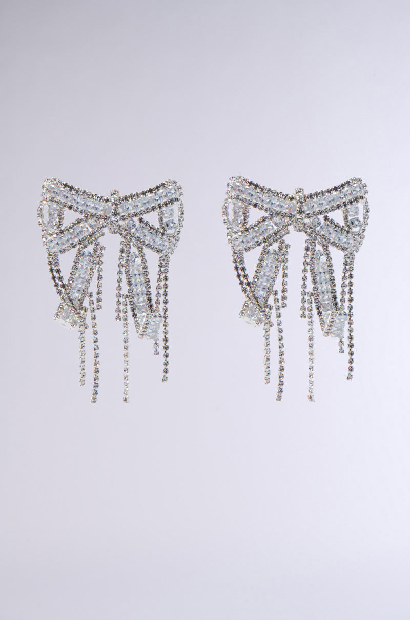 The WITH A BOW EARRING features sparkling rhinestone embellishments and dangling strands, shown against a plain light gray background.