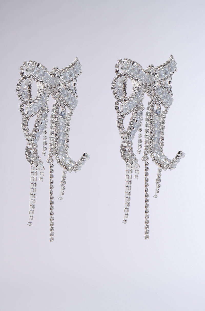 WITH A BOW EARRING: Sparkling silver bow earrings feature rhinestone accents and long, dangling strands, shown against a plain light gray background—a chic statement for any occasion.
