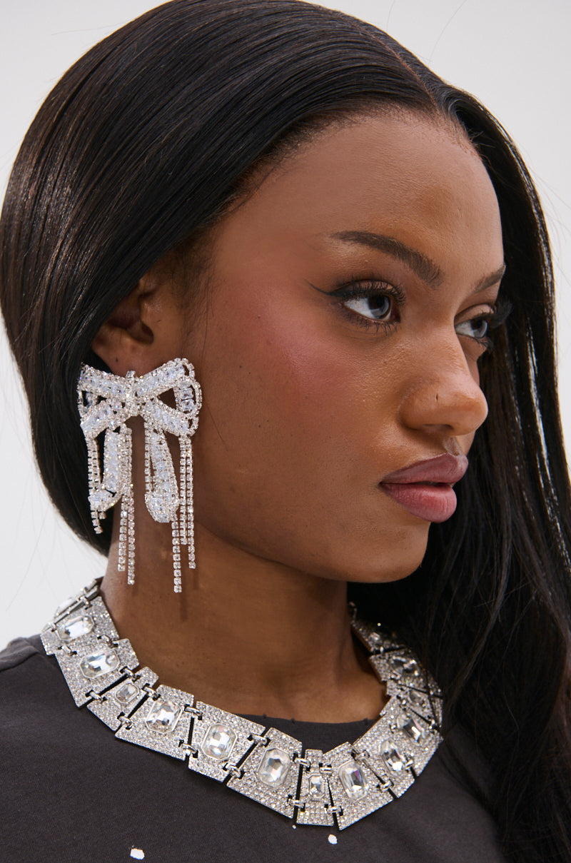 A woman with long dark hair wears the WITH A BOW EARRING, a large sparkly accessory adorned with rhinestones, along with a chunky geometric silver necklace. She has winged eyeliner and a neutral expression, facing to the right.