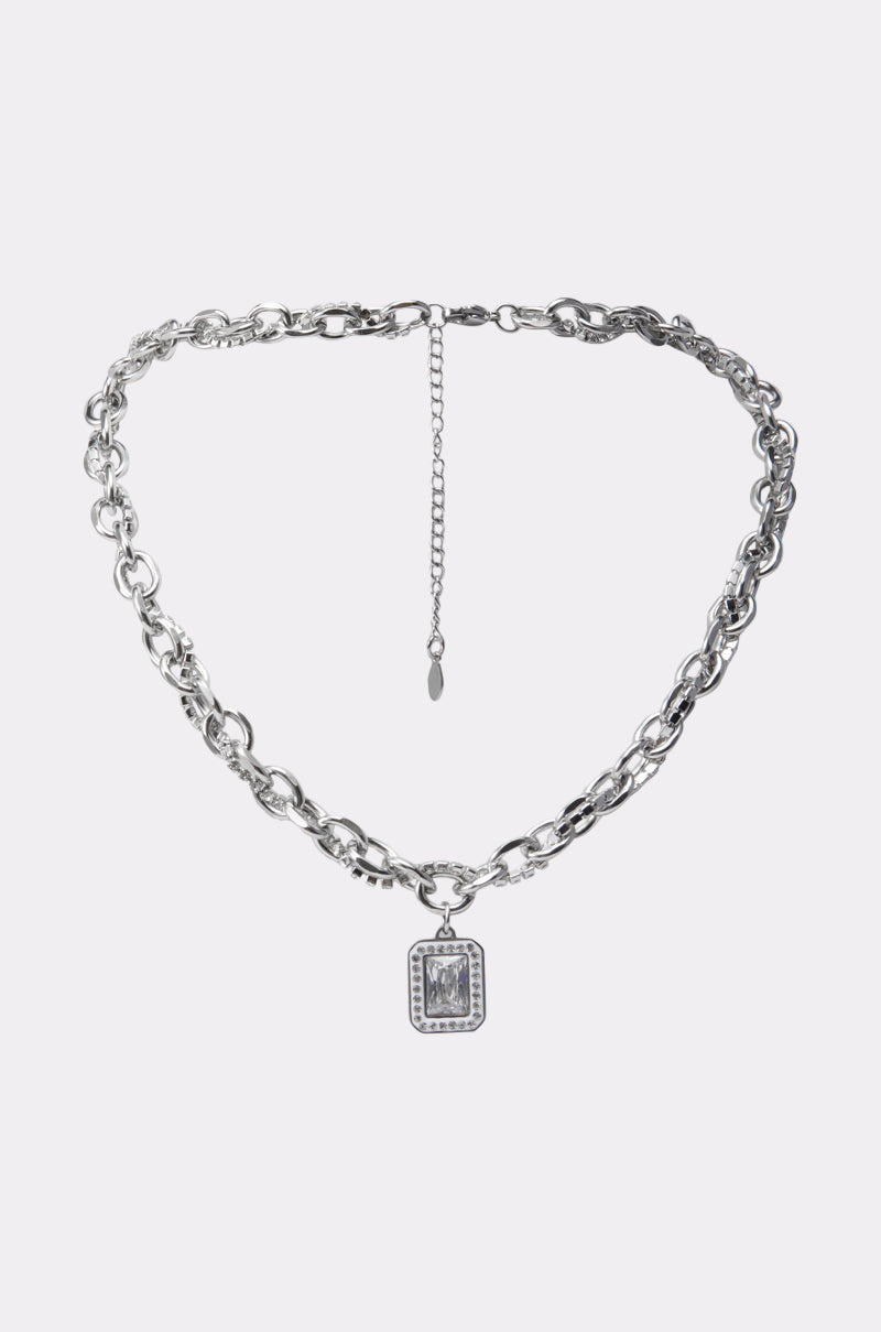 The SHES EXPENSIVE SILVER NECKLACE features mixed links, an adjustable clasp, and a rectangular pendant with a clear gem framed by small stones. Its chunky curb chain stands out against a plain white background. 1