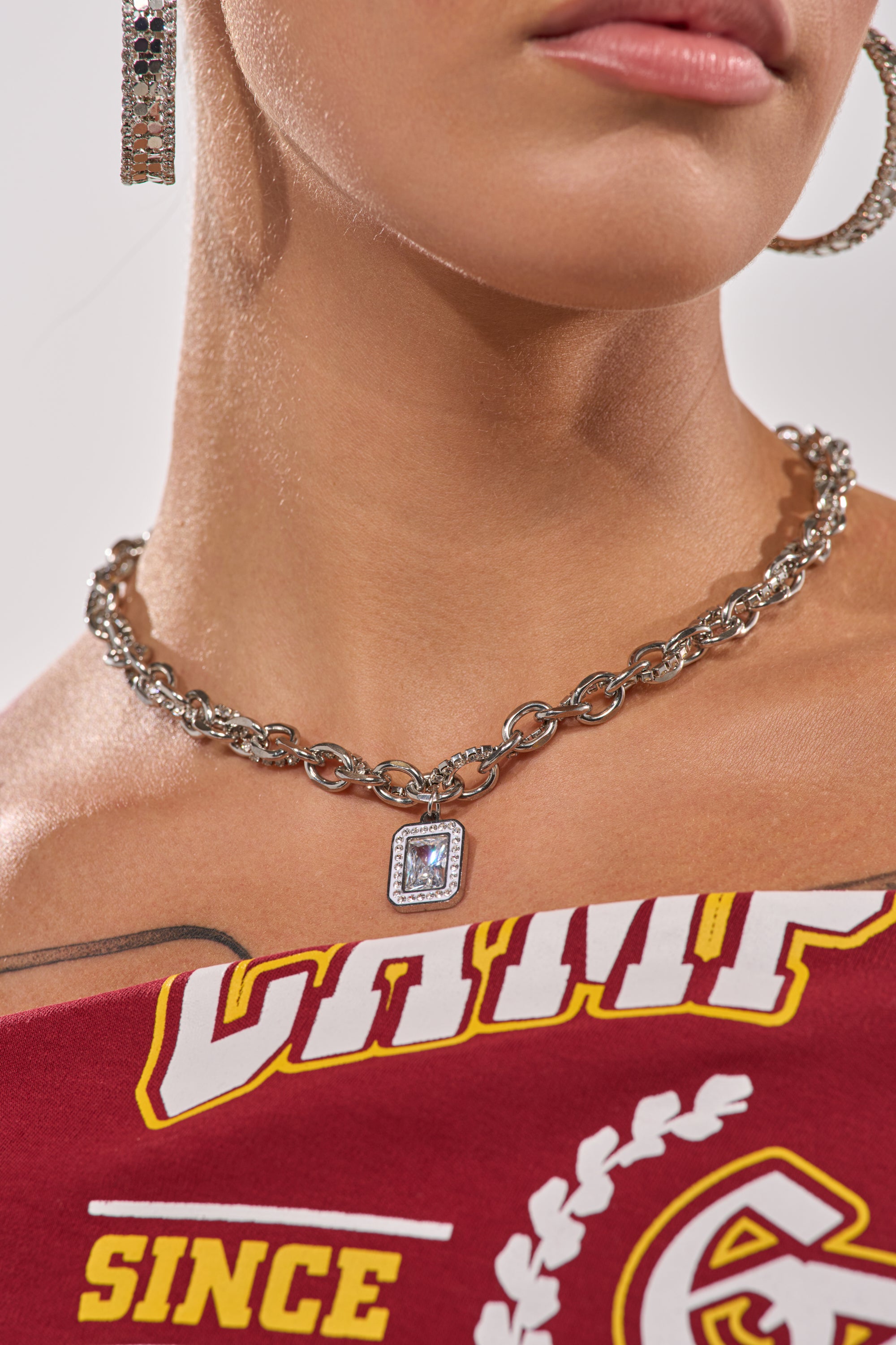 Close-up of a person wearing the SHES EXPENSIVE SILVER NECKLACE, featuring a chunky curb chain and rectangular pendant, paired with sparkly hoop earrings and a red top with yellow and white "CAMP" and "SINCE" lettering. 2