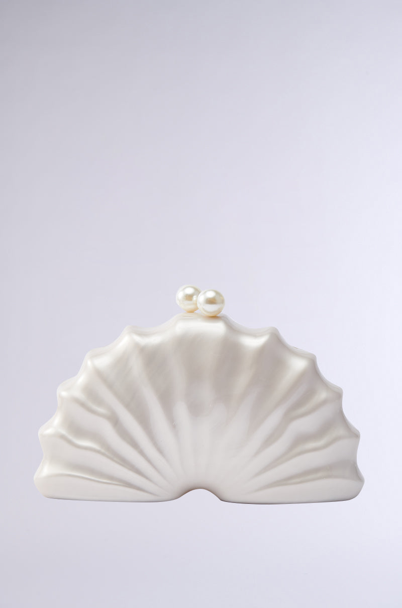 The IN TULUM CLUTCH is a cream-colored, shell-shaped purse featuring a ridged texture and two pearl-like clasps on top, displayed against a plain light gray background. 0