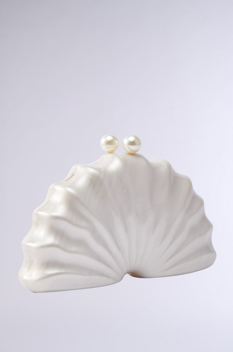 The IN TULUM CLUTCH is a white, shell-shaped purse featuring a smooth wavy texture and twin pearl-like clasps, shown against a plain light gray background. 2