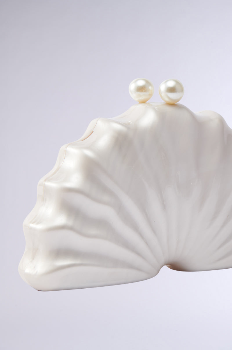 The IN TULUM CLUTCH is a white, clam-shaped purse with a smooth, glossy finish and two round pearl clasps, shown against a plain light gray background. 3