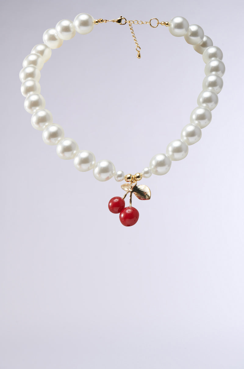The CHERRY ON TOP NECKLACE features large round faux pearls, a gold-tone chain clasp, and a cherry pendant with two red beads and gold leaves at the center.