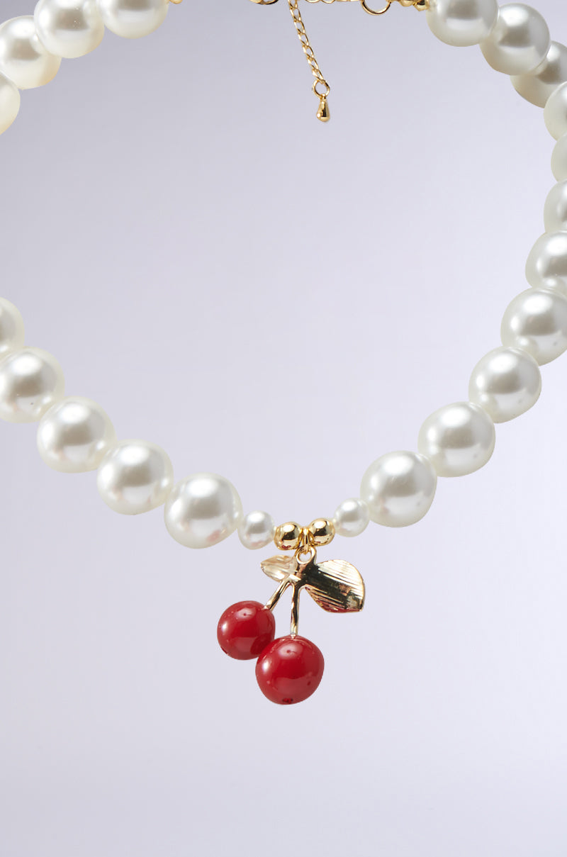 The CHERRY ON TOP NECKLACE features faux pearls with a gold charm of two glossy red cherries and a gold leaf, shown on a light gray background.