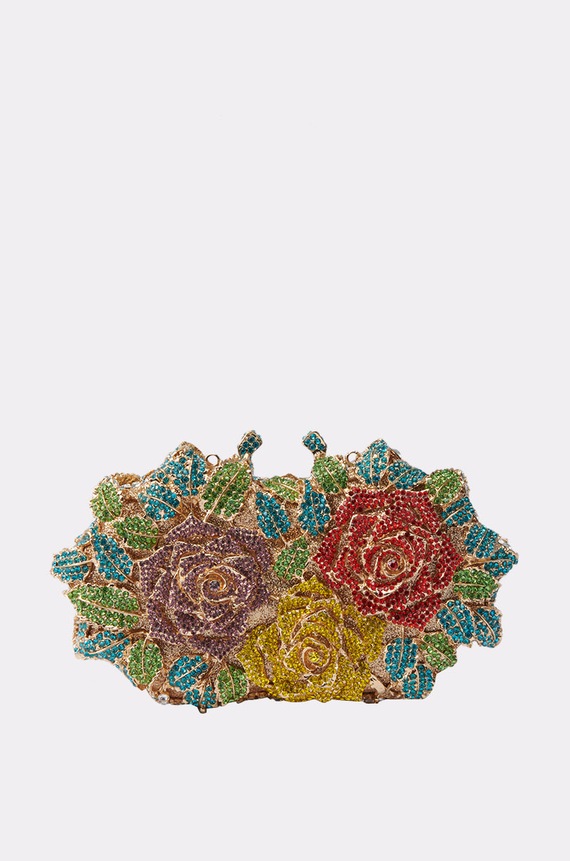 The IN THE GARDEN BLING BAG is a decorative clutch purse with a sculptural silhouette, sparkling beaded roses in purple, red, and yellow, and green leaves set against a plain white background.
