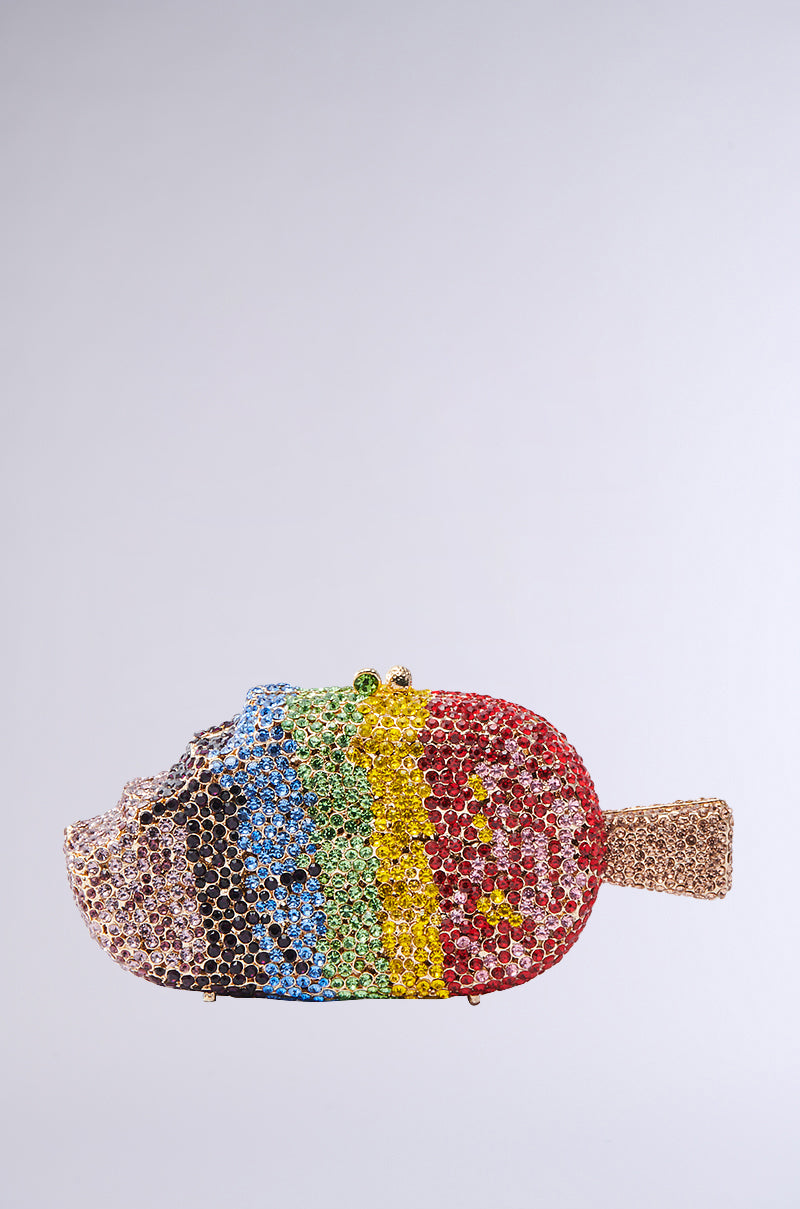 The LICK IT BLING BAG is a sparkling fish-shaped clutch adorned with vibrant rhinestones in brown, blue, green, yellow, and red stripes, finished with a gold tail and clasp. Displayed against a plain light gray background.
