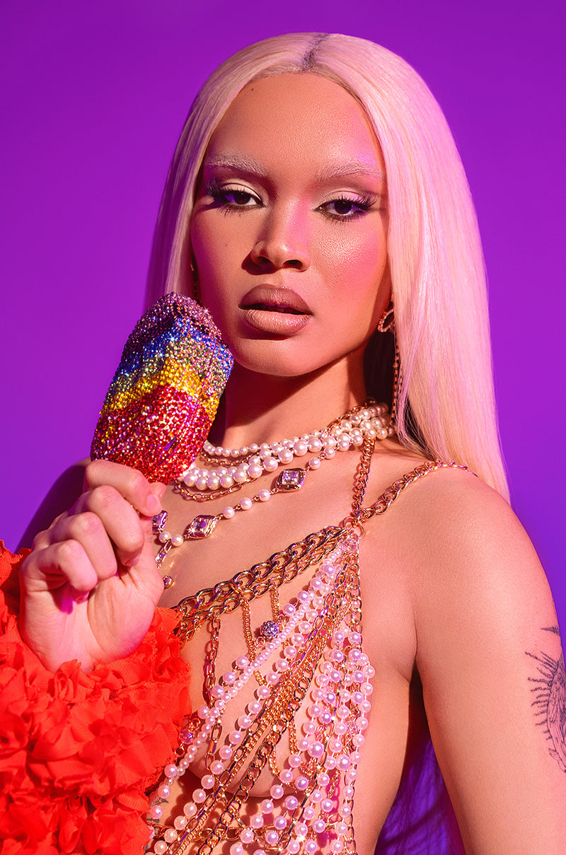 With long platinum blonde hair and dramatic makeup, a person holds a gem-studded popsicle, wears layered pearls and chains, and carries the LICK IT BLING BAG—a metallic hardshell accessory—against a vivid purple backdrop with red ruffled fabric.
