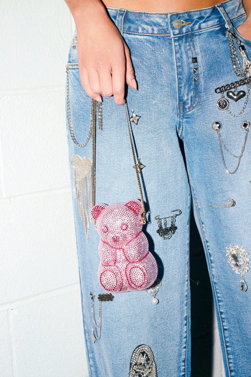 A person in blue jeans with metallic chains and charms holds the YUMMY GUMMY BLING BAG by its chainlink strap. The photo is cropped to show only their lower torso and hand.