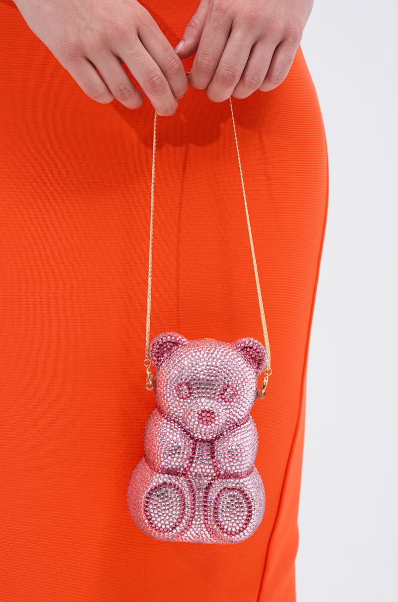 A person in an orange dress holds the YUMMY GUMMY BLING BAG—a pink rhinestone teddy bear-shaped bag with a gold chainlink strap.