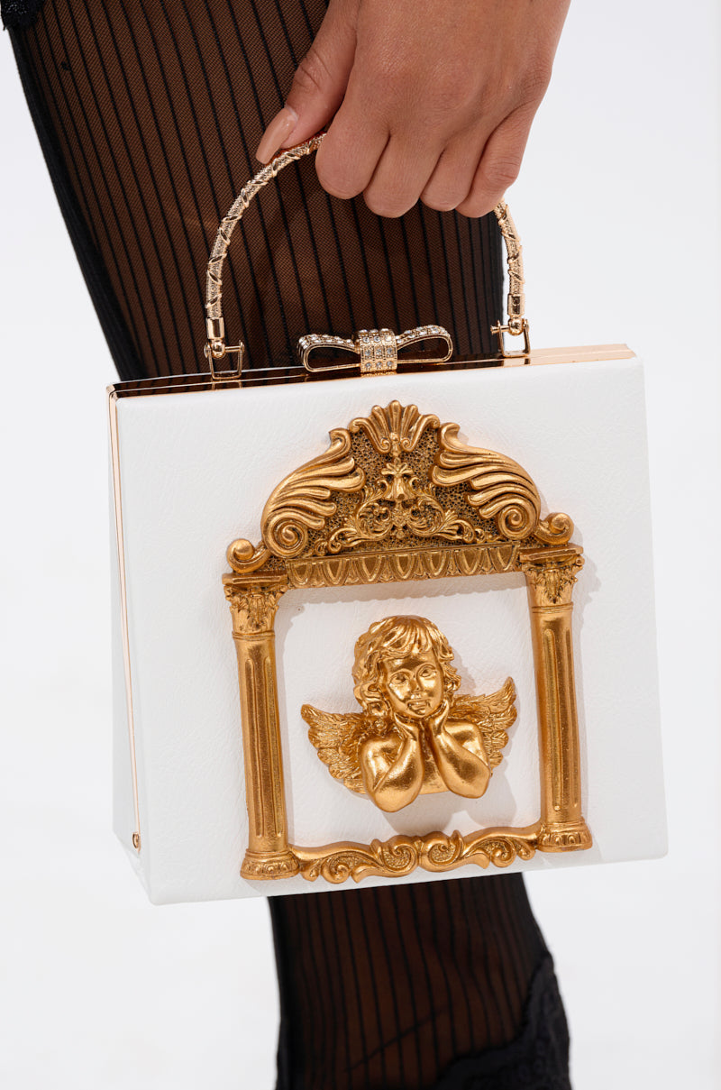 A person holds the IM AN ANGEL PURSE, a white box handbag with a gold ornate frame, angel design, and decorative gold bow handle. They wear black sheer pleated stockings, highlighting the purse’s baroque clutch style.