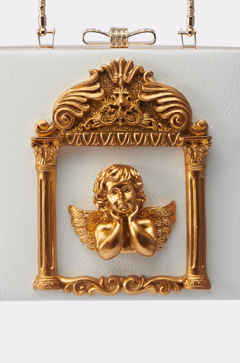 The IM AN ANGEL PURSE features a golden cherub with wings resting its face on its hands, framed by an ornate gold arch with columns, embodying vintage glamour against a white textured backdrop.