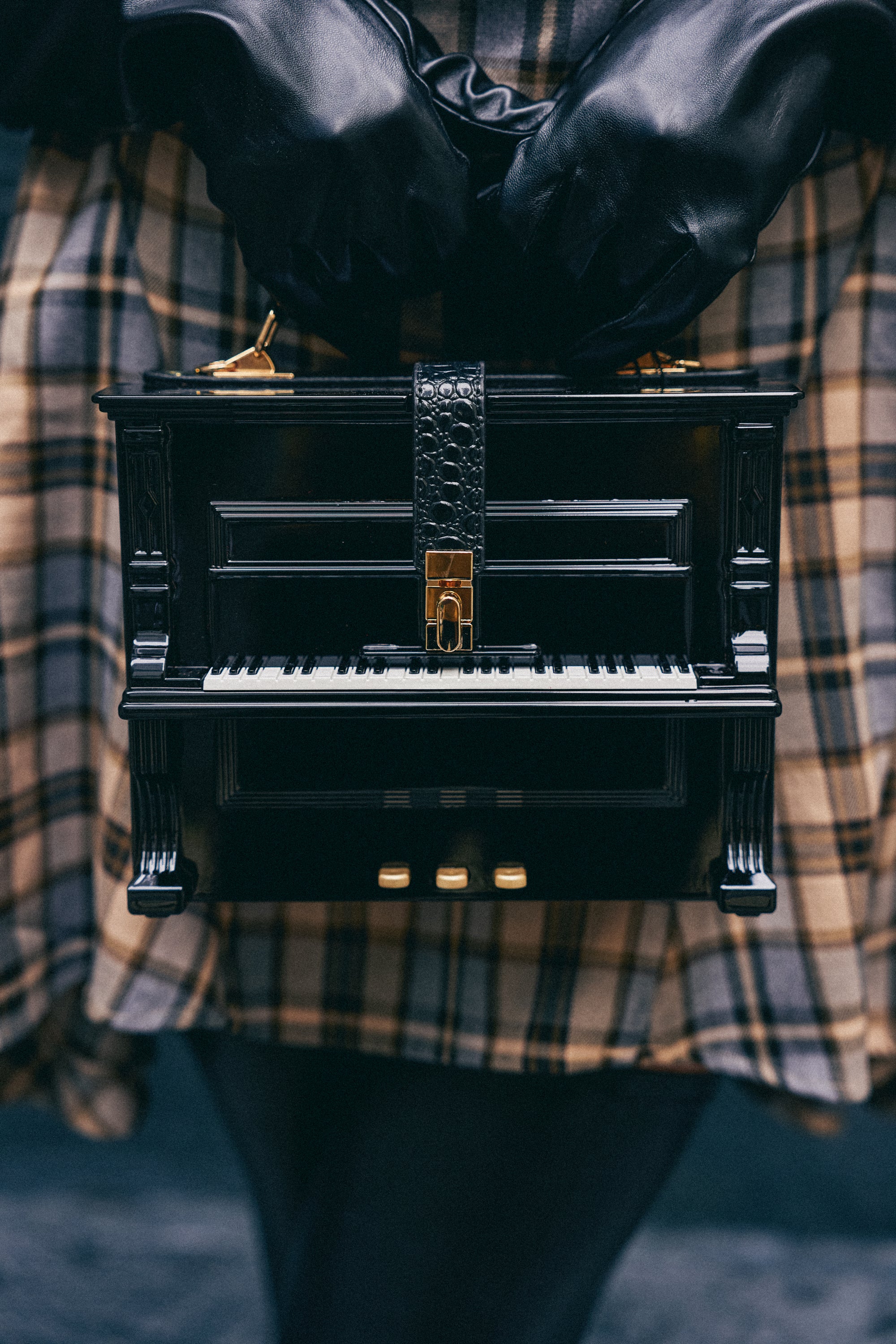 A person in plaid holds the PLAY ME PIANO PURSE, a black novelty bag shaped like a grand piano, while wearing black gloves.
