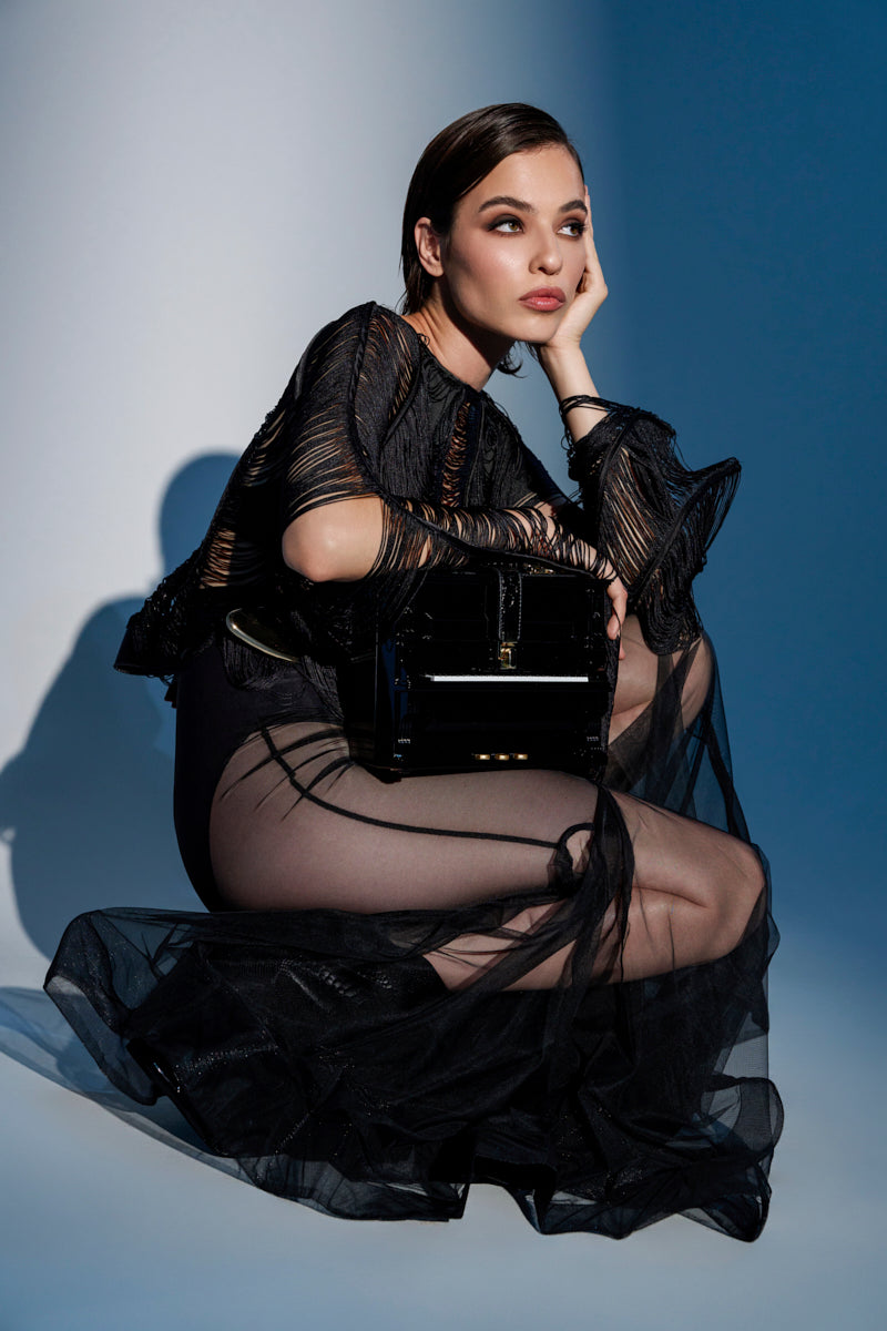A woman in a sheer black dress sits on the floor, one hand on her cheek, looking thoughtful. She holds the PLAY ME PIANO PURSE, its shiny surface highlighted by dramatic lighting that casts bold shadows behind her.