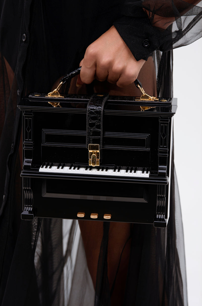 A person in a sheer black outfit holds the PLAY ME PIANO PURSE, designed as a miniature black piano with white and black keys, gold accents, crocodile pattern, and a top handle.
