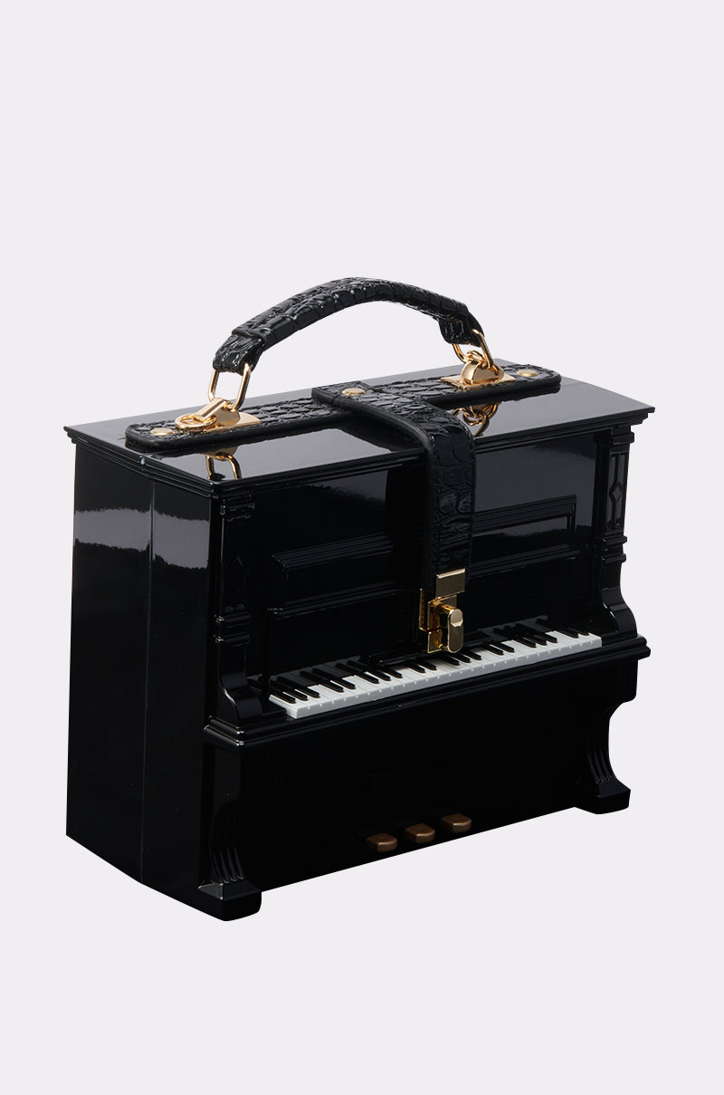The PLAY ME PIANO PURSE is designed to look like a black upright piano with keys, pedals, and a crocodile pattern handle with gold hardware, shown against a plain background.