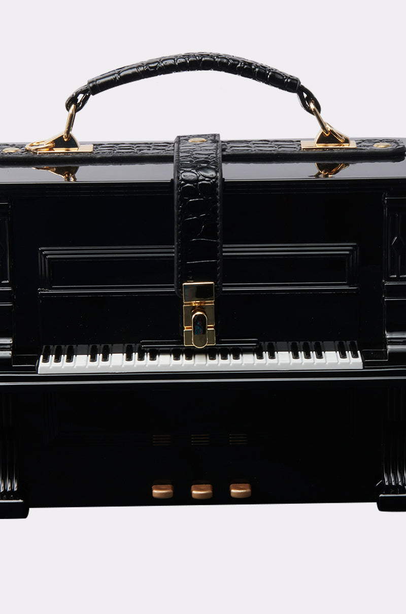 The PLAY ME PIANO PURSE is a black bag styled as a grand piano, with white and black keys, a crocodile pattern handle, and gold hardware accents including the central clasp.
