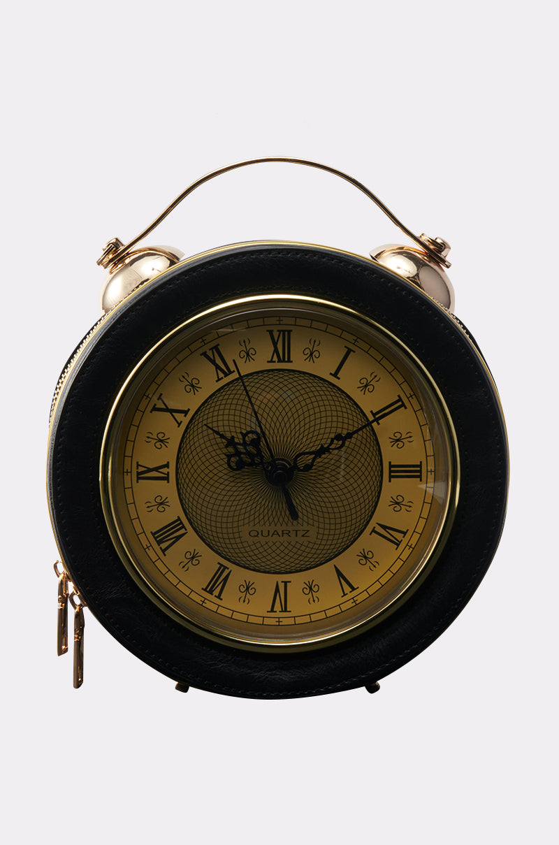 The WHATS THE TIME PURSE is a vintage-style round alarm clock featuring a gold face, black Roman numerals, ornate black hands, a leather-like black frame, and top handle—resembling a classic clock purse—set against a plain light background.