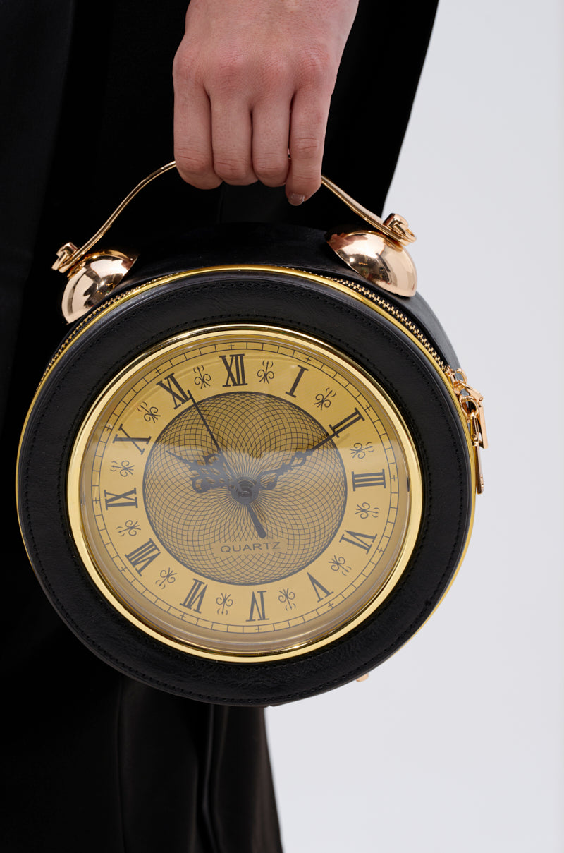 A person holds the WHATS THE TIME PURSE, a round black and gold bag styled like a vintage alarm clock with Roman numerals and clock hands on the front.