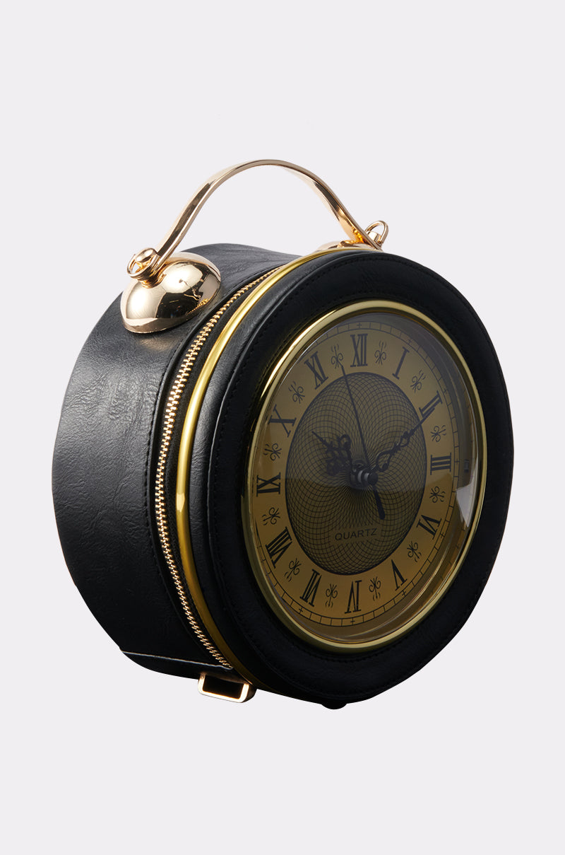 The WHATS THE TIME PURSE features a vintage clock face with Roman numerals on a round, black faux leather bag with a gold zipper and top handle, blending timeless charm with modern style.
